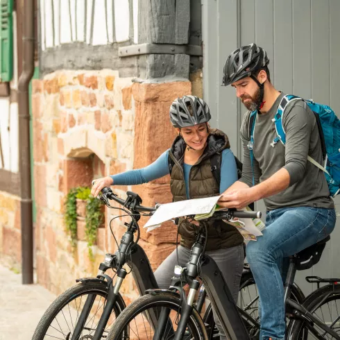 Ein Mann und eine Frau mit Fahrradhelm und zwei Fahrrädern schauen gemeinsam in eine Radkarte, im Hintergrund ein Fachwerkhaus des Ortes Steinweiler im Pfälzer Bergland